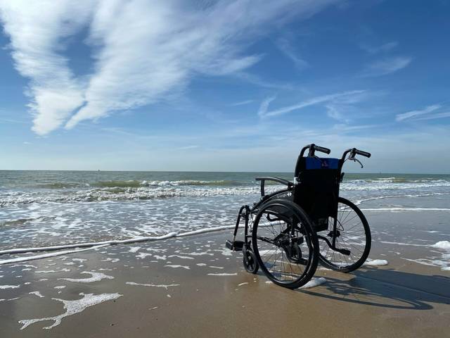 Empty wheelchair on a sandy beach near gentle waves, under a blue sky with wispy clouds.
