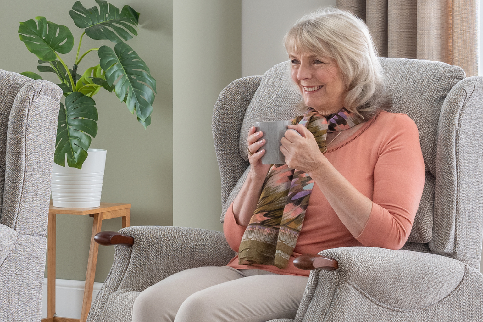 Smiling woman with a scarf sits in a cozy armchair, holding a mug. Indoor plants are arranged in the background.