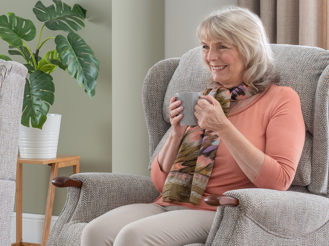 Smiling woman with a scarf sits in a cozy armchair, holding a mug. Indoor plants are arranged in the background.