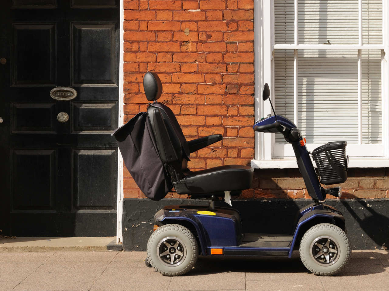 A mobility scooter parked on a sidewalk next to a brick building with a black door and white window shutters.
