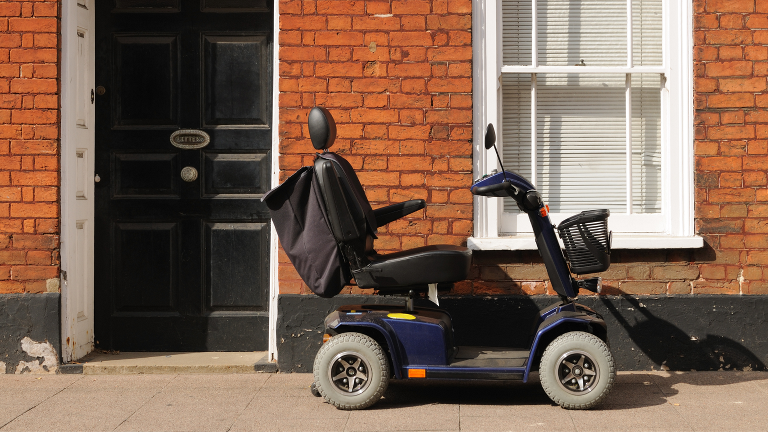 A mobility scooter parked on a sidewalk next to a brick building with a black door and white window shutters.