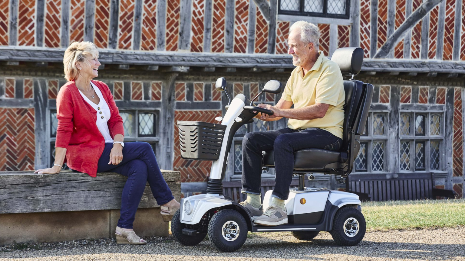 An elderly man using a mobility scooter converses with a woman sitting on a bench outside a rustic building.