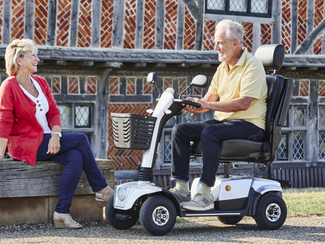An elderly man using a mobility scooter converses with a woman sitting on a bench outside a rustic building.