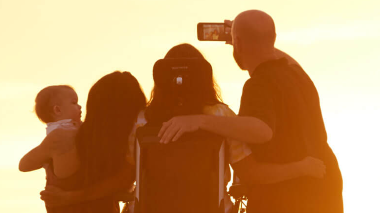 A group of four people, including a child, take a selfie at sunset. One is in a wheelchair, creating a silhouette against the glowing sky.