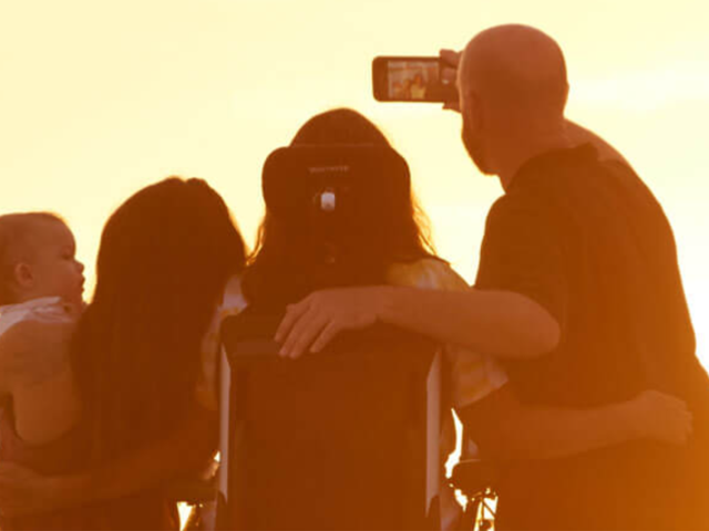 A group of four people, including a child, take a selfie at sunset. One is in a wheelchair, creating a silhouette against the glowing sky.