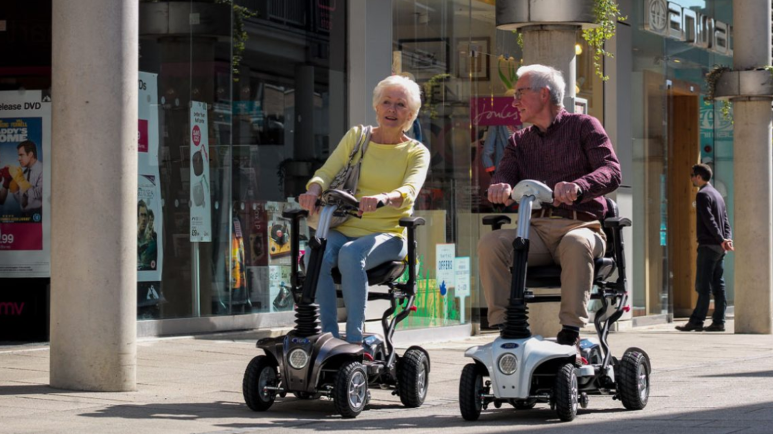 Two elderly people ride mobility scooters, smiling and talking, against a backdrop of shopfronts on a sunny day.