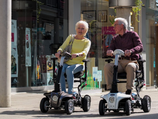 Two elderly people ride mobility scooters, smiling and talking, against a backdrop of shopfronts on a sunny day.
