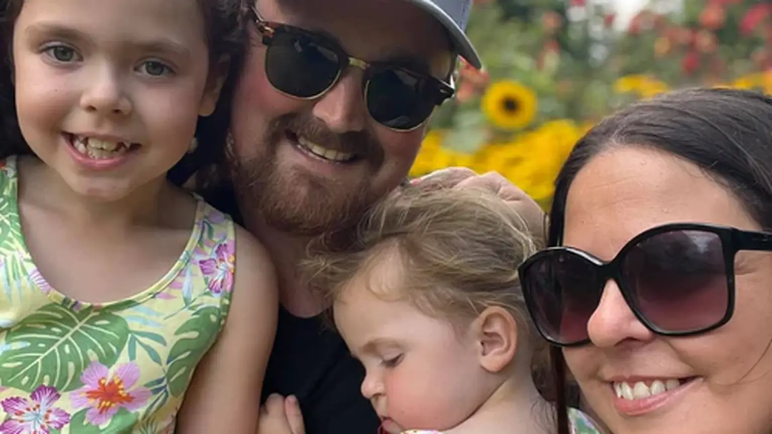 A family of four poses outdoors, surrounded by sunflowers. The two children wear matching floral dresses, and the adults wear sunglasses.