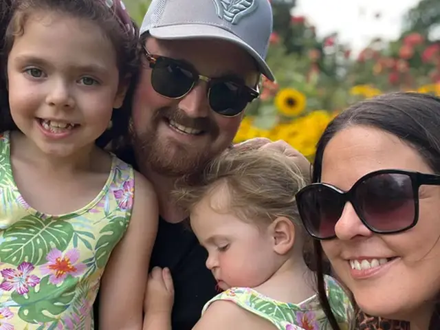 A family of four poses outdoors, surrounded by sunflowers. The two children wear matching floral dresses, and the adults wear sunglasses.