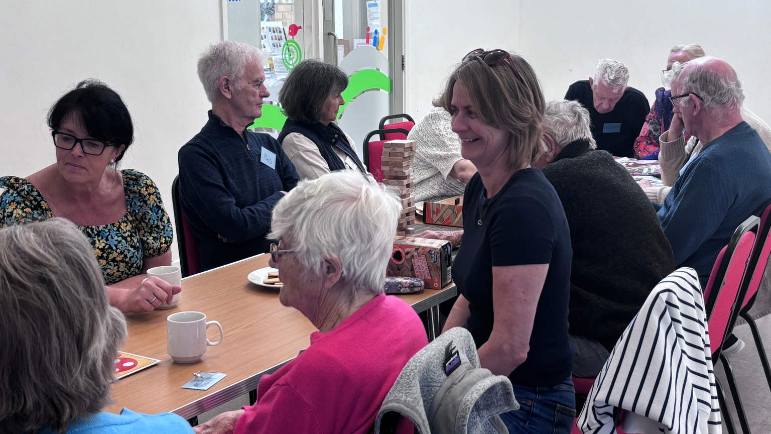 A group of people sitting around a table, chatting and enjoying drinks in a community setting, with games visible on the table.