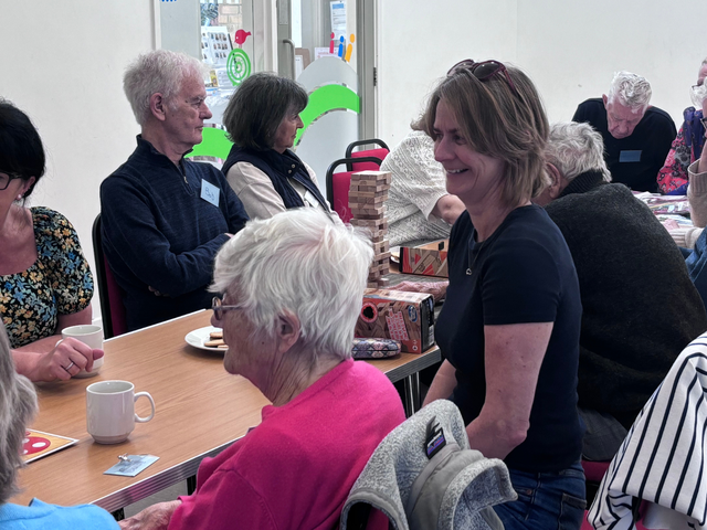 A group of people sitting around a table, chatting and enjoying drinks in a community setting, with games visible on the table.