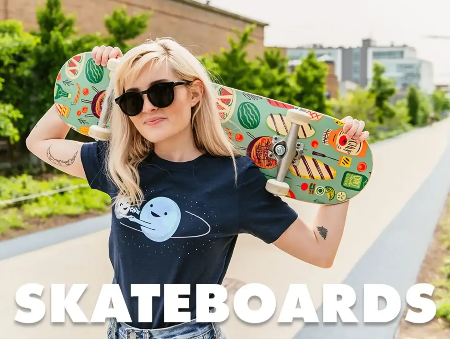 Person carrying colorful planet print skateboard outdoors wearing teal t-shirt with 'Skateboards' text.