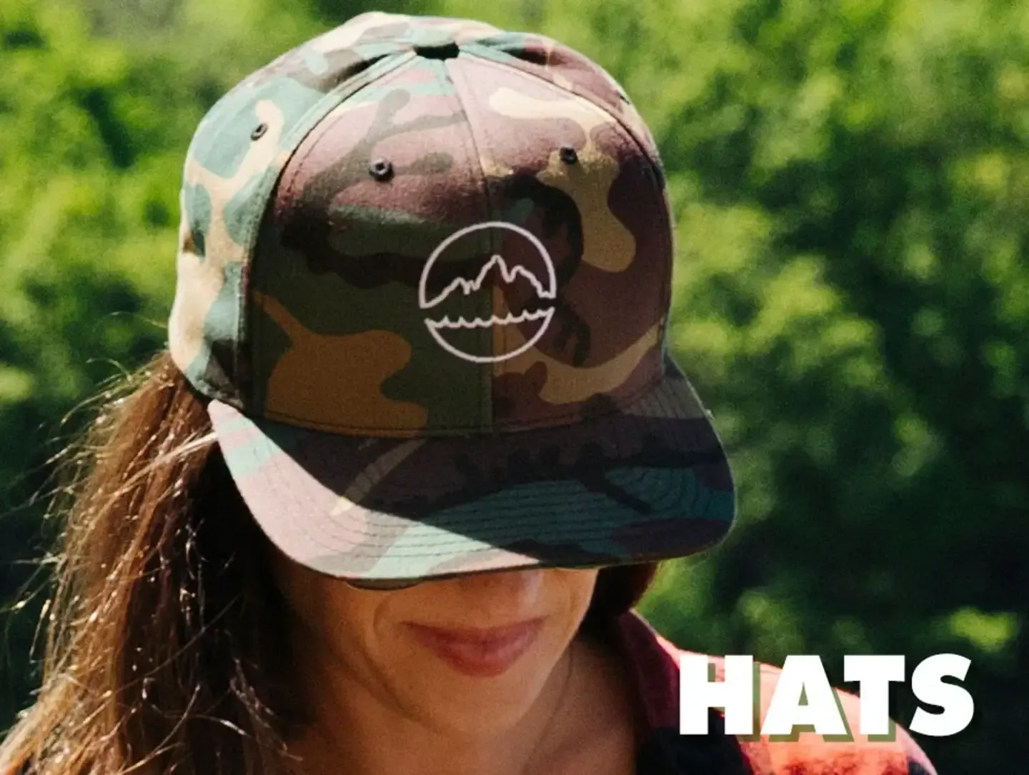 Smiling person in camouflage baseball hat with mountain logo against brick wall with 'Hats' text.