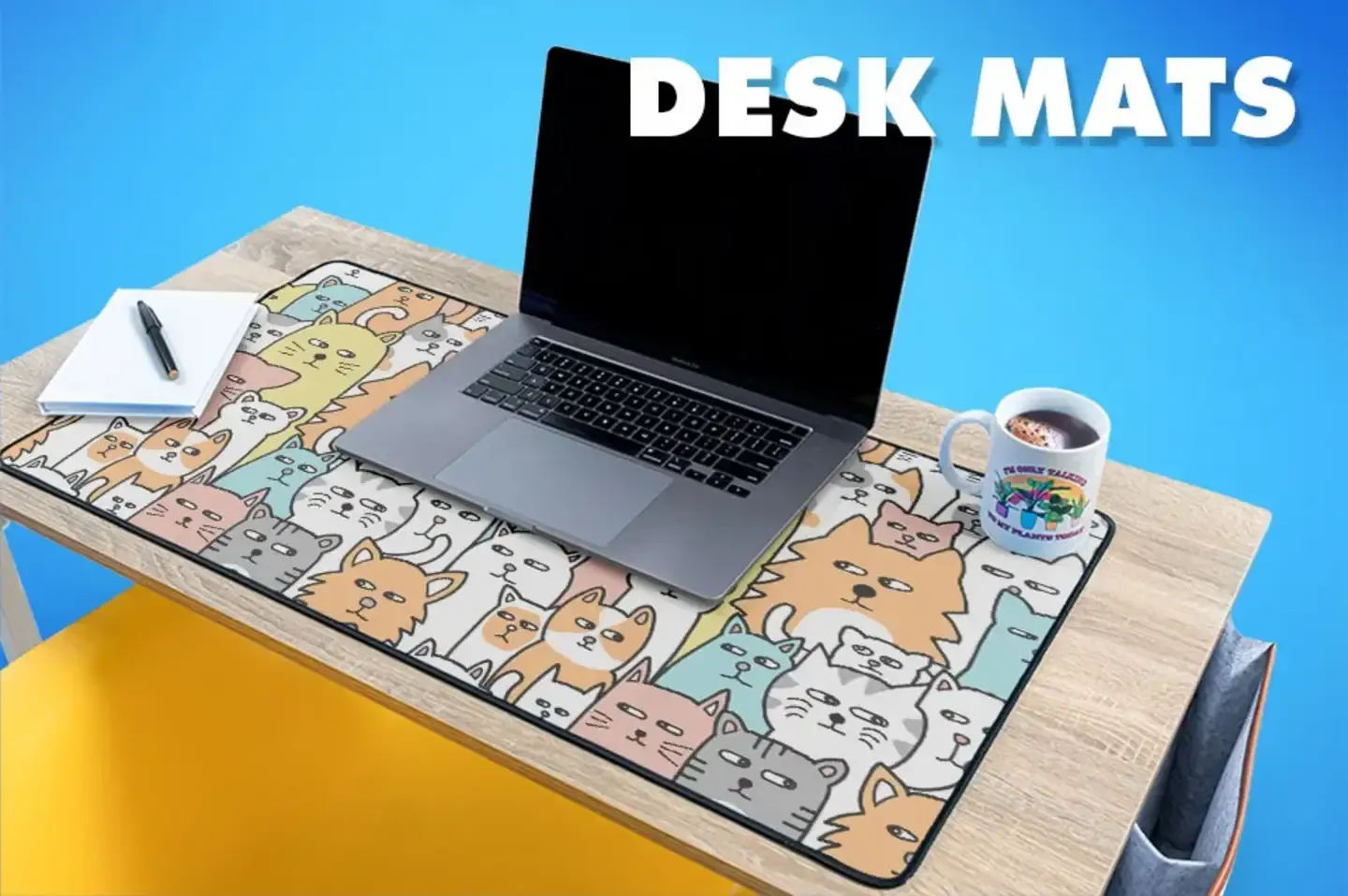 Colorful cat illustration desk mat under open laptop with coffee mug on yellow desk with 'Desk Mats' text.