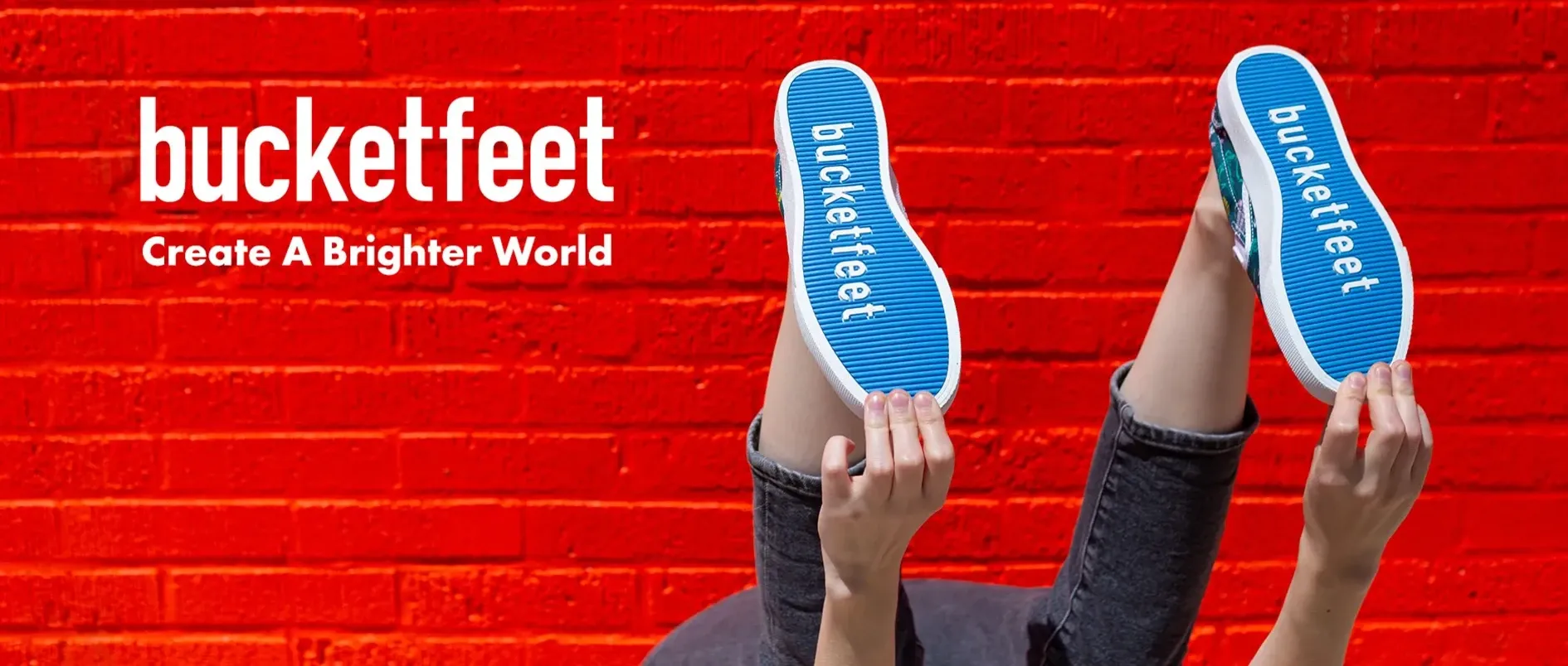 Hands holding up a pair of Bucketfeet slip-on shoes against a red brick wall, showing the blue soles