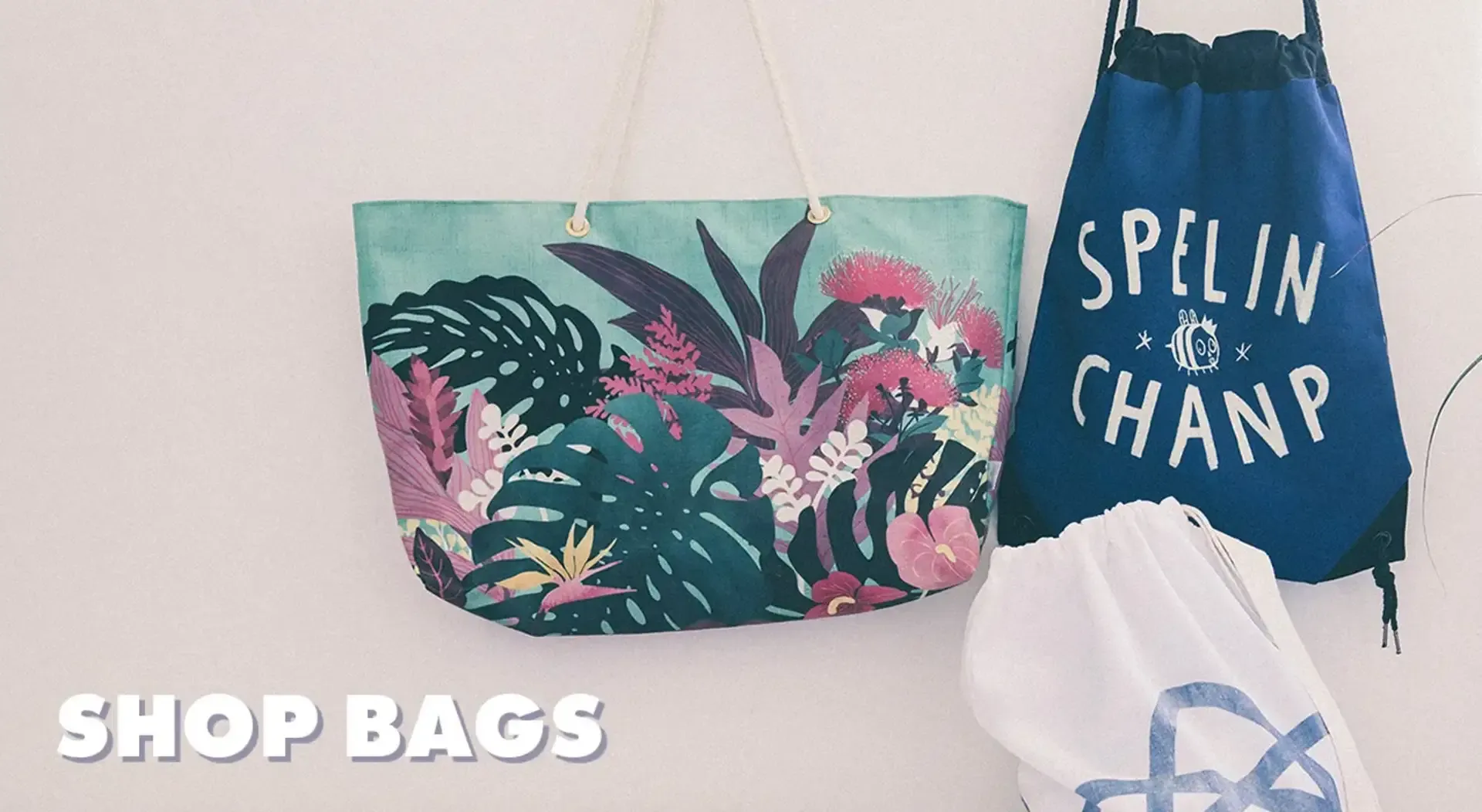 Tropical print tote bag and 'Spelin Champ' navy drawstring bag hanging on white wall with 'Shop Bags' text.