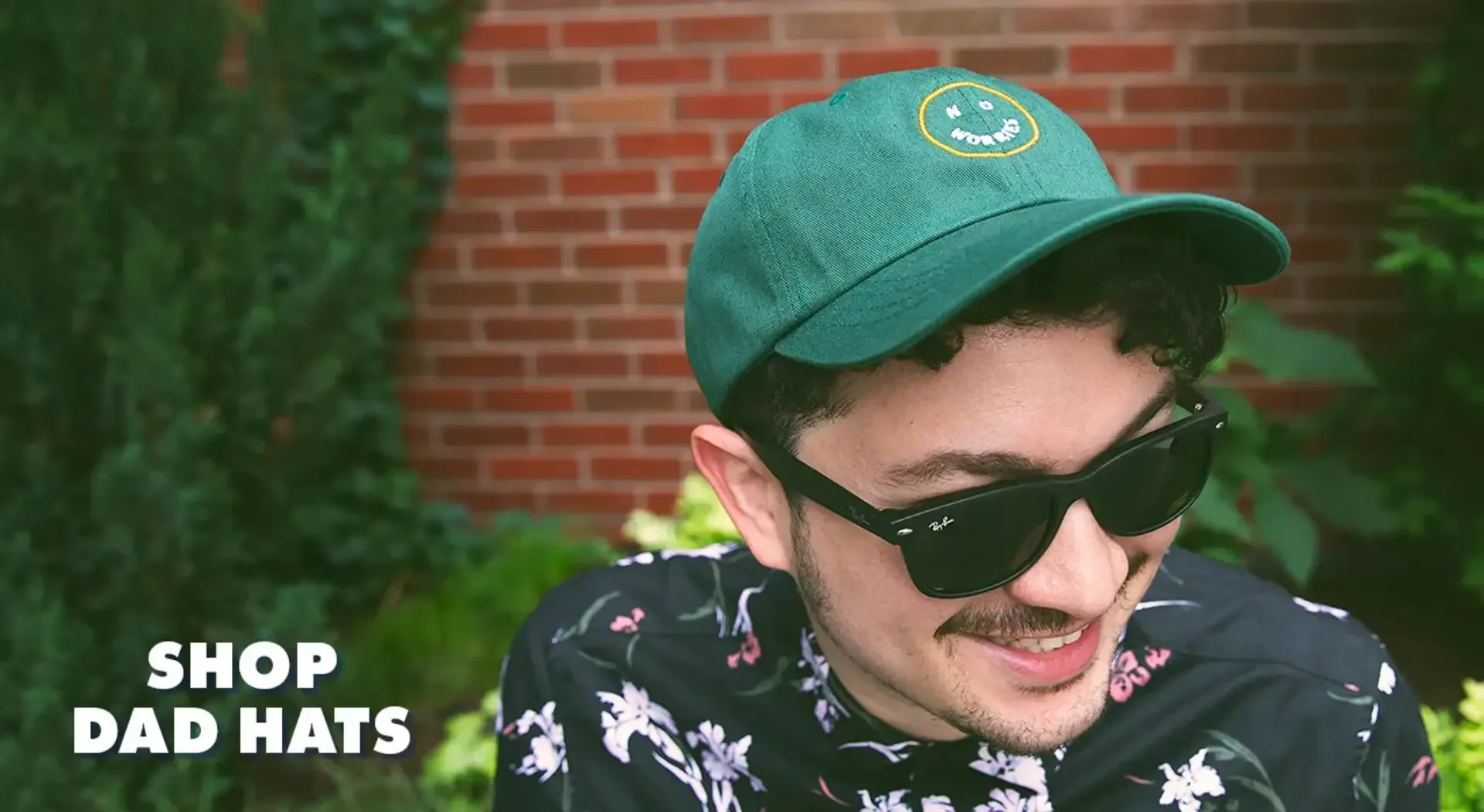 Smiling person in sunglasses wearing teal dad hat with embroidered smiley patch against brick wall.