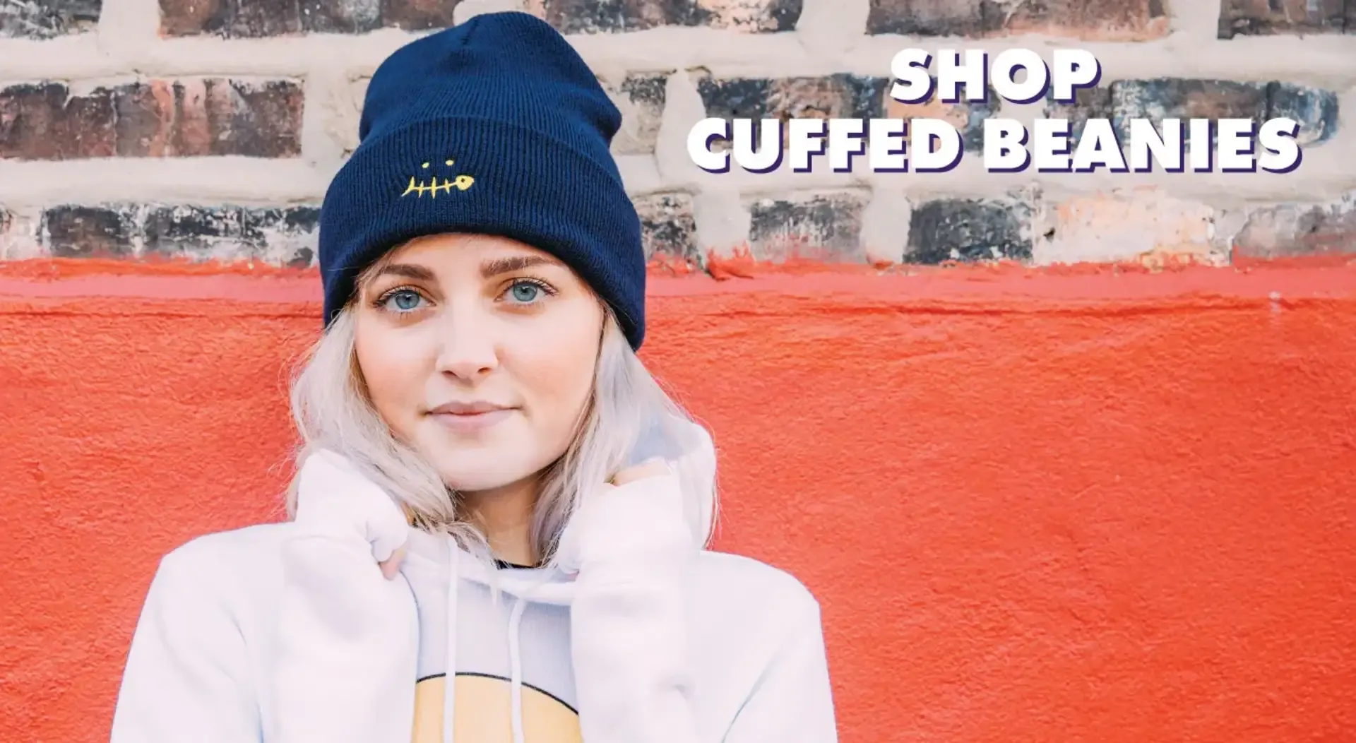 Blonde person wearing navy cuffed beanie with embroidered face design against red brick wall.