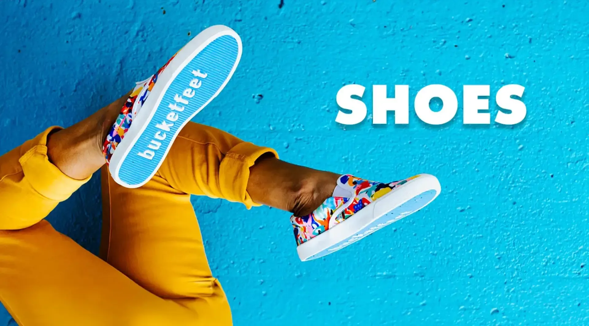Woman's legs in yellow pants wearing colorful Bucketfeet sneakers against bright blue painted wall background.
