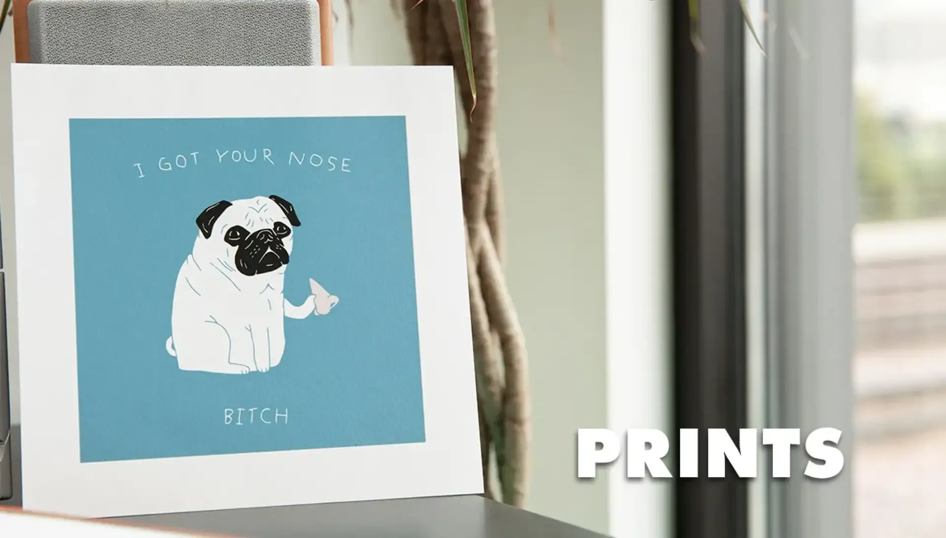 Framed 'I Got Your Nose' pug illustration print on shelf in bright room labelled 'Prints'.