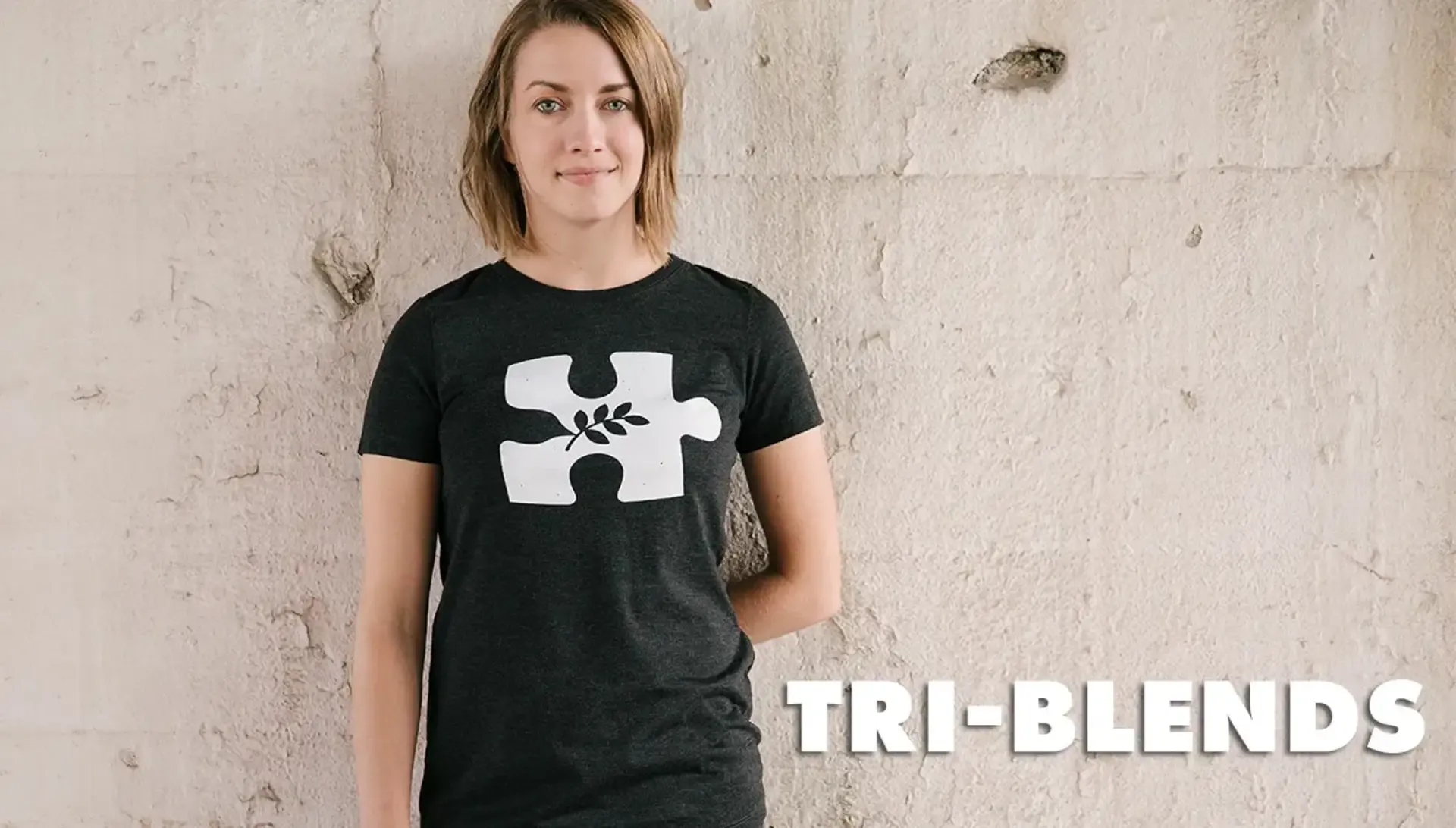 Woman in dark heather women's tri-blend t-shirt with white Threadless puzzle piece logo on concrete backdrop.