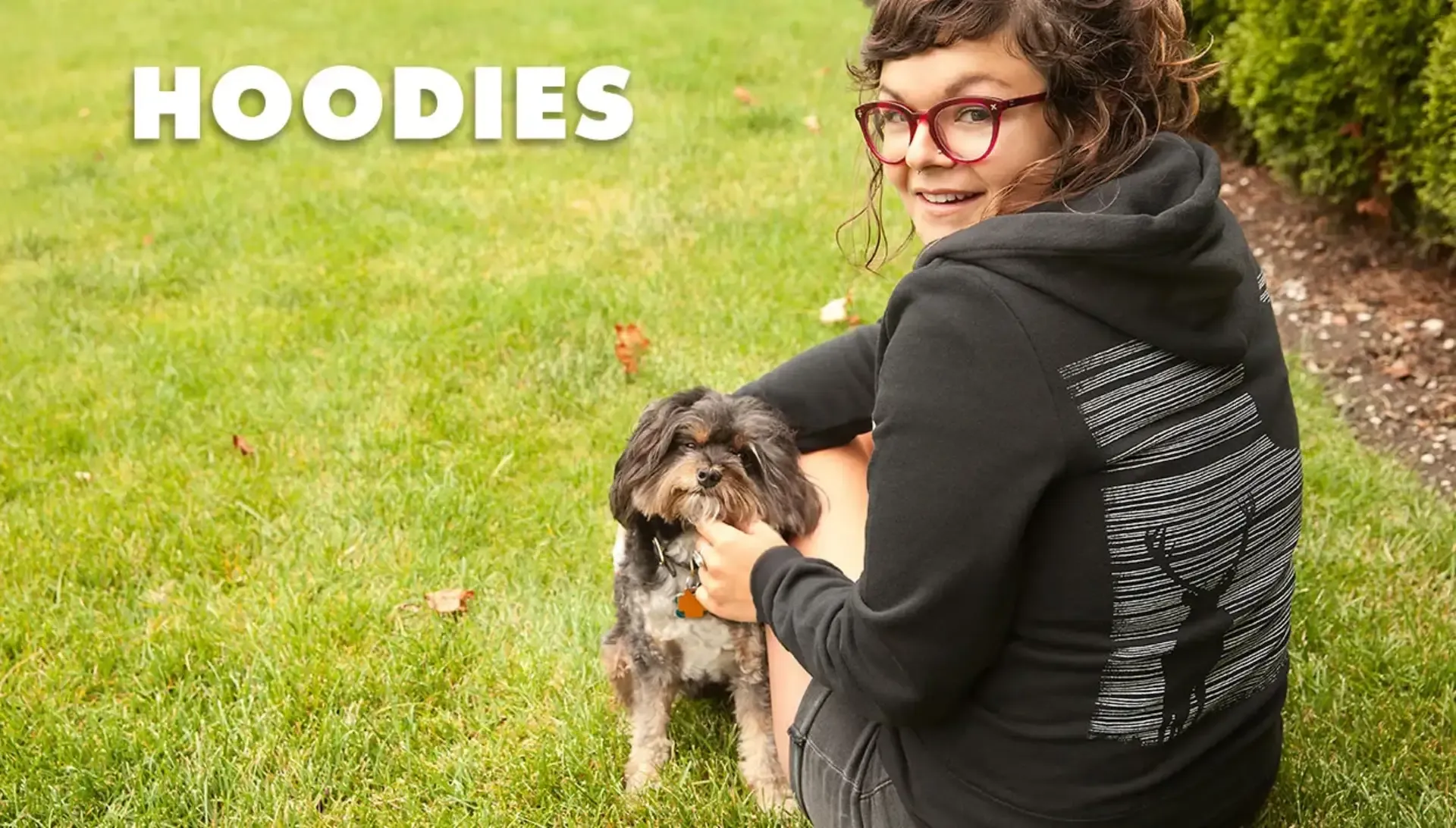 Woman in glasses holding small dog on grass, wearing black women's hoodie with white deer forest graphic.