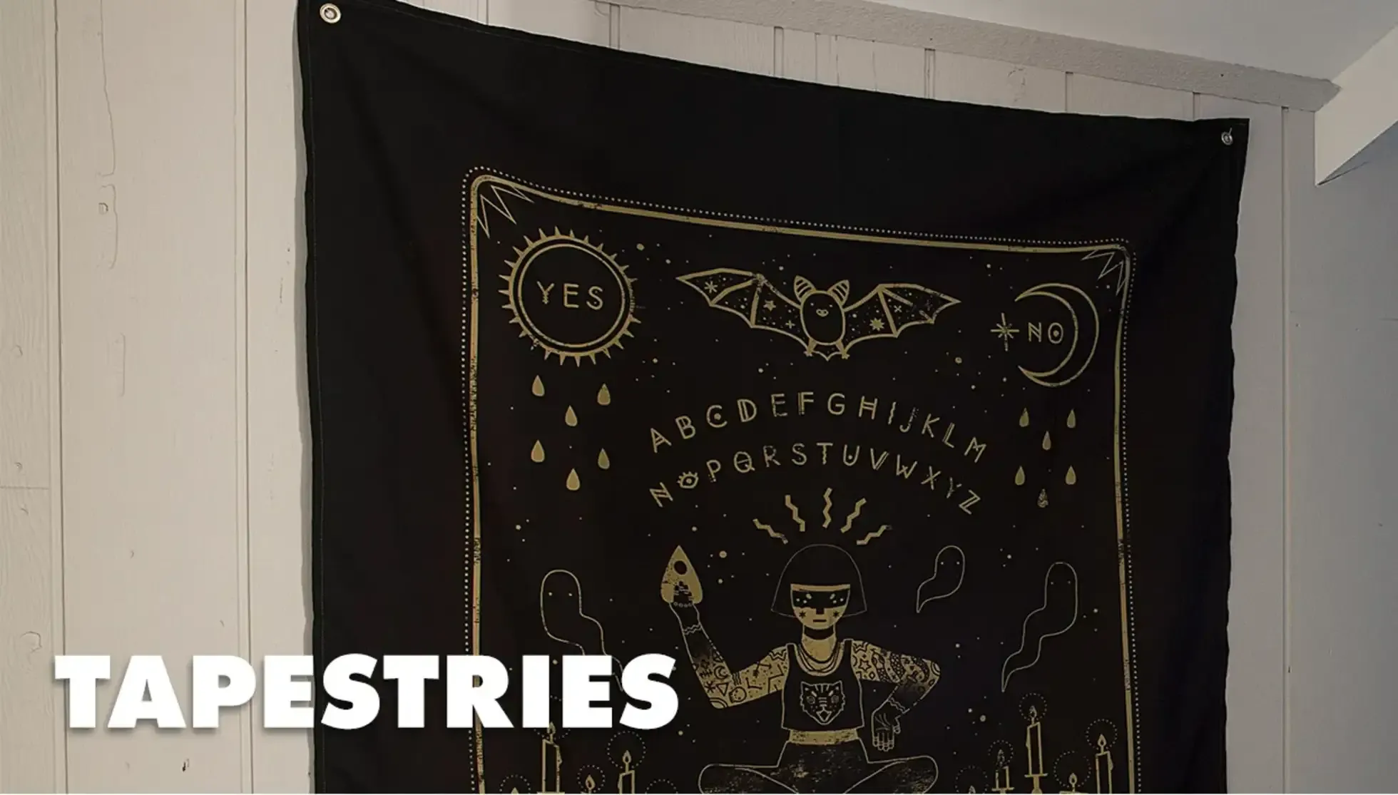 Black ouija board alphabet tapestry hanging on white wall labelled 'Tapestries'.