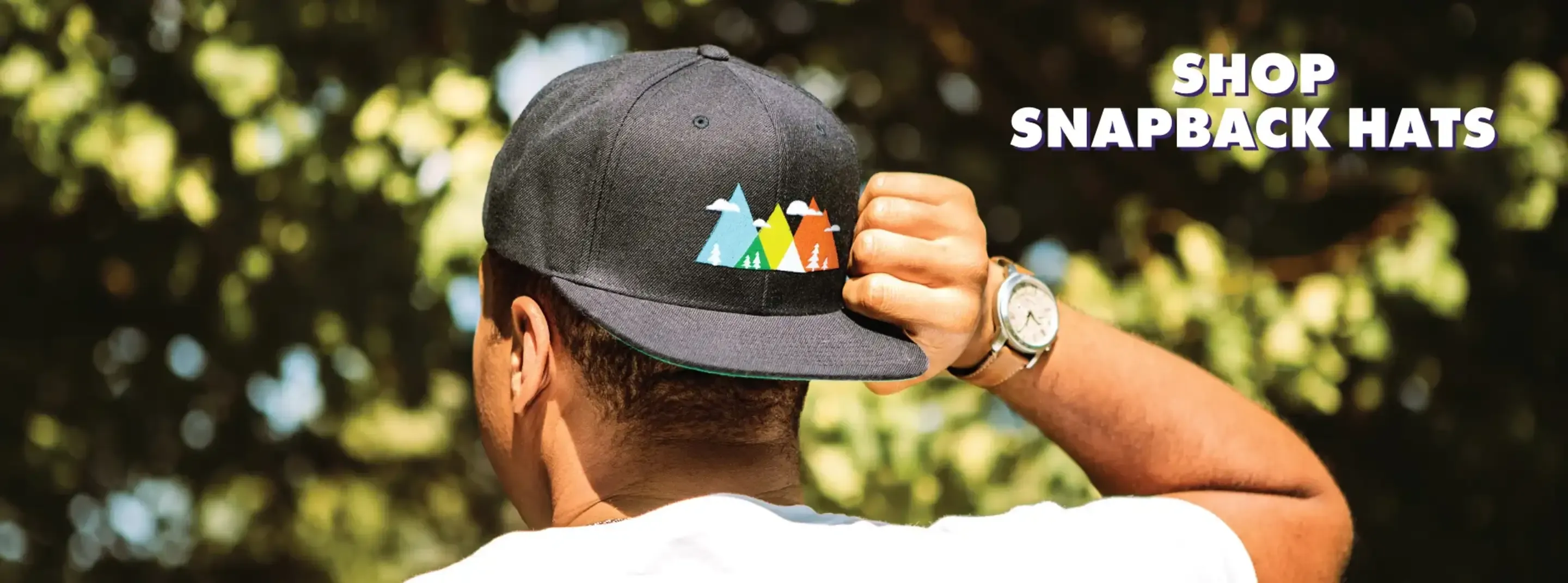 Person wearing black snapback hat with mountain graphic, viewed from behind against tree foliage.