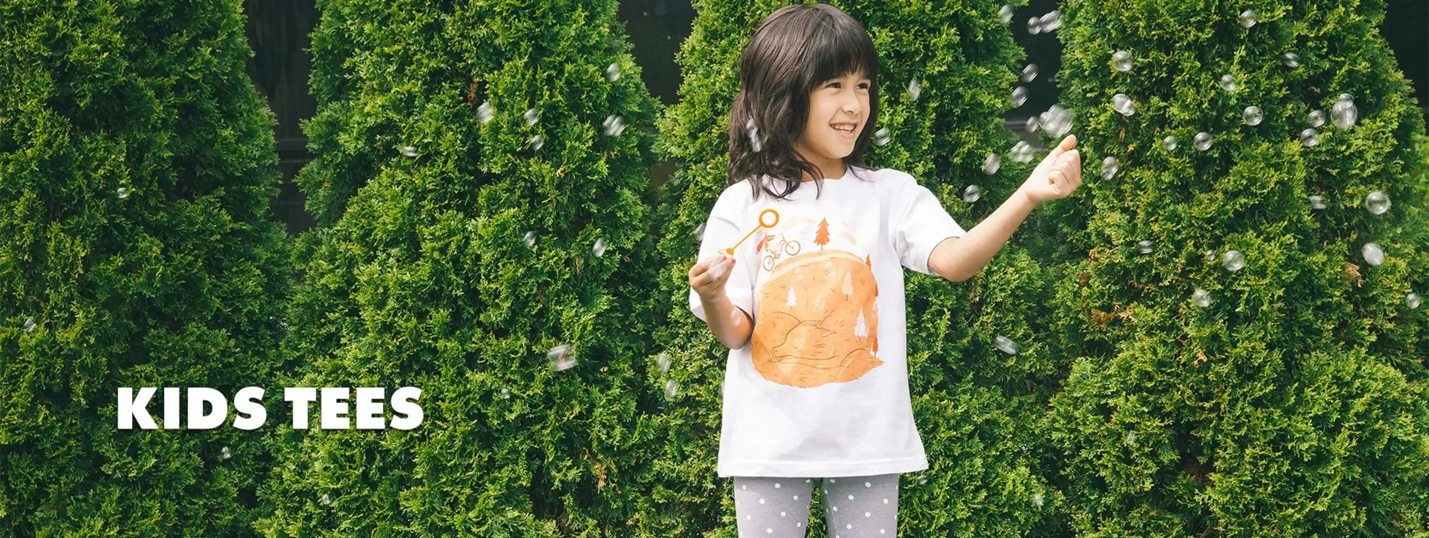 Child blowing bubbles in white kids t-shirt with sandwich illustration against green hedge background, labelled 'Kids Tees'.
