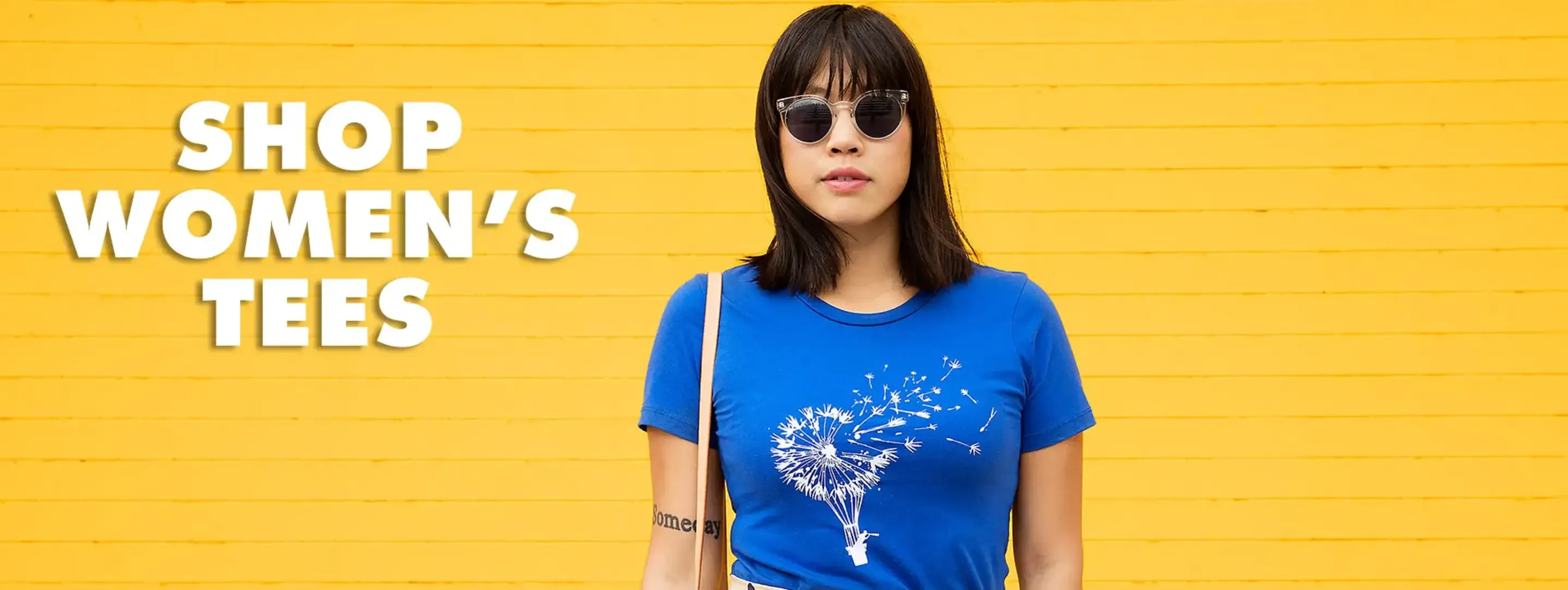 Woman in sunglasses wearing blue women's t-shirt with white dandelion seeds graphic on yellow wall backdrop.