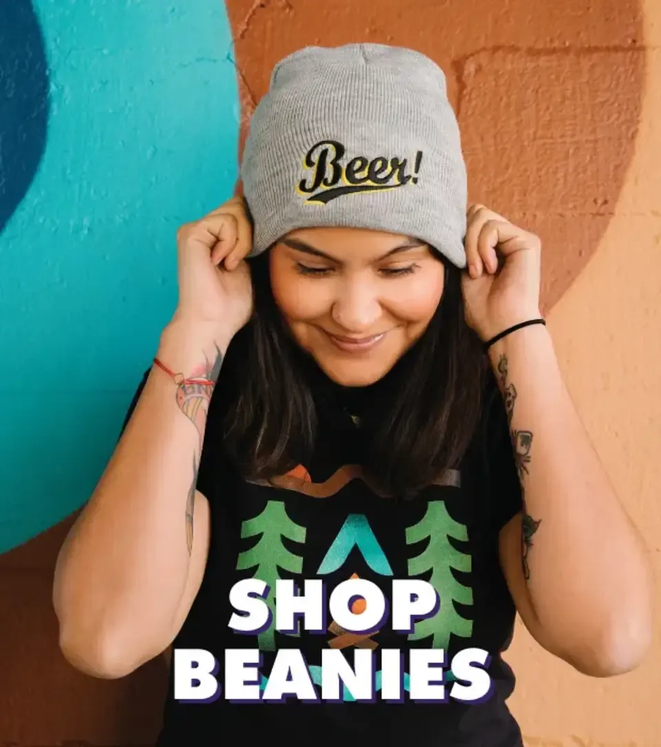 Smiling person wearing cream beanie with 'Beer' embroidery and tree print t-shirt against teal wall.