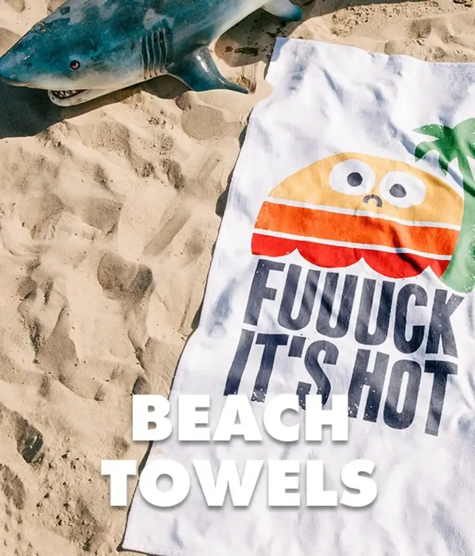 Burger character beach towel on sand near shark fin prop with 'Fuuuck It's Hot' design and 'Beach Towels' text.