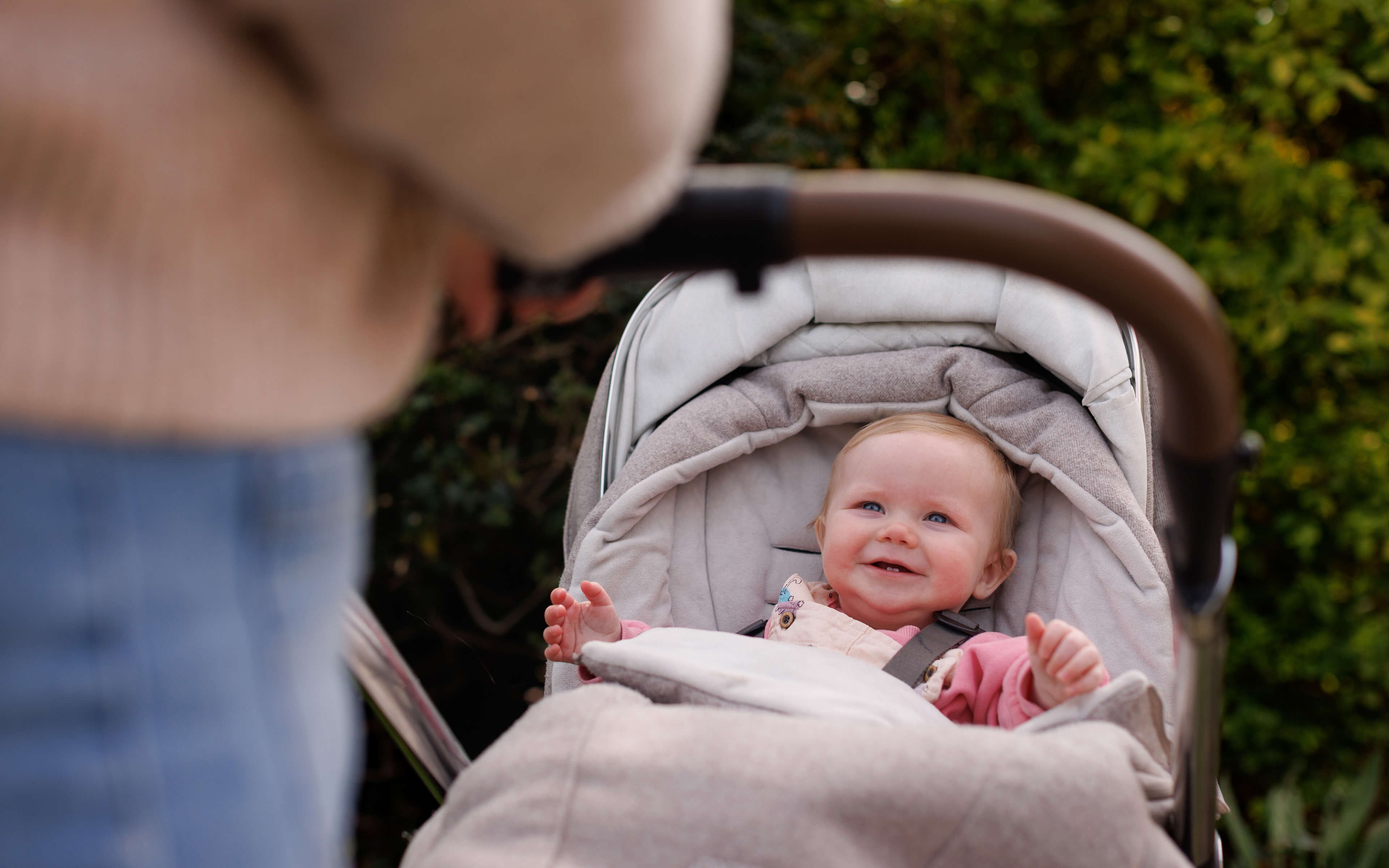 A smiling baby in a stroller looks up at an adult, surrounded by green foliage in the background