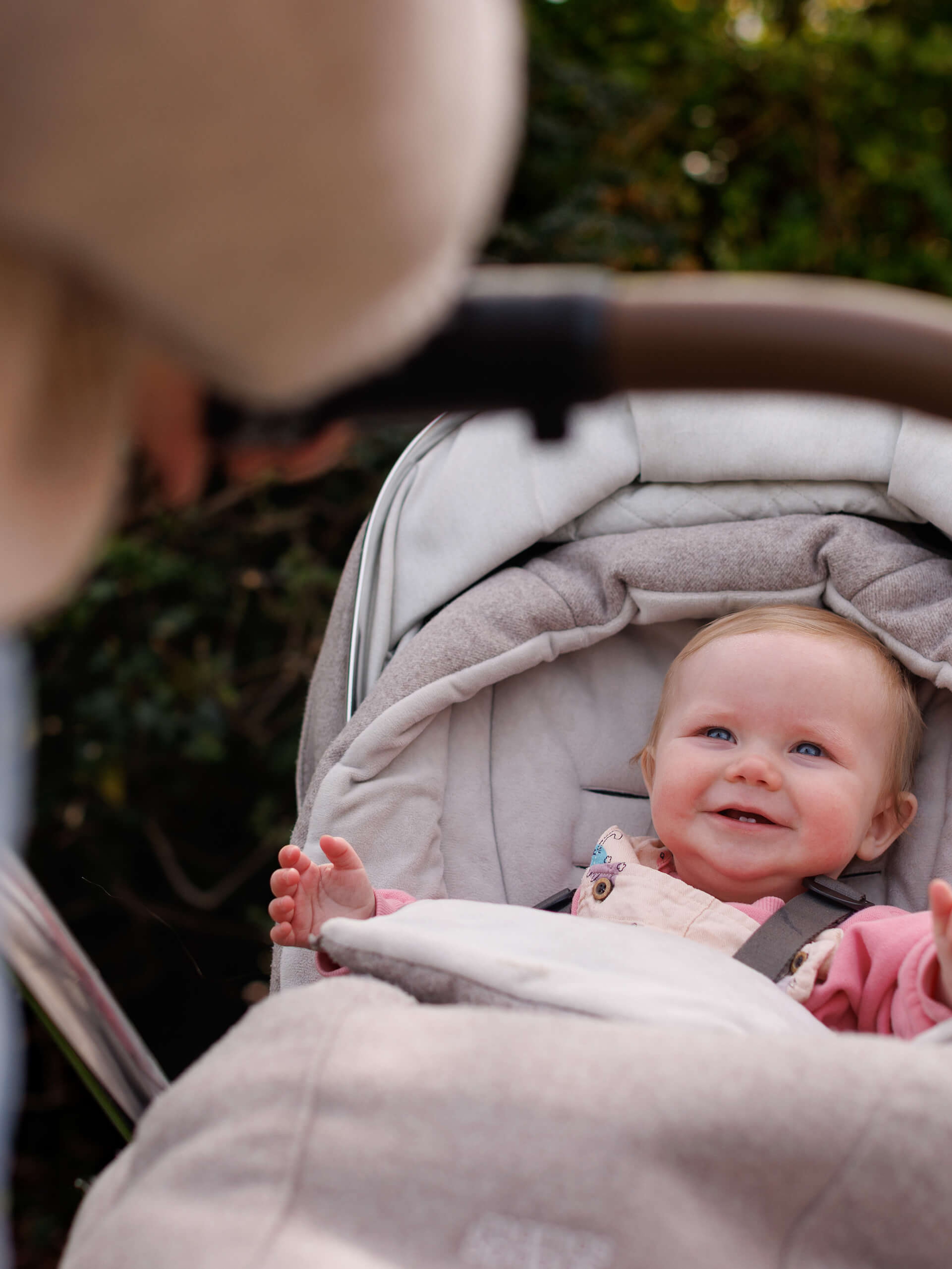 A smiling baby in a stroller looks up at an adult, surrounded by green foliage in the background