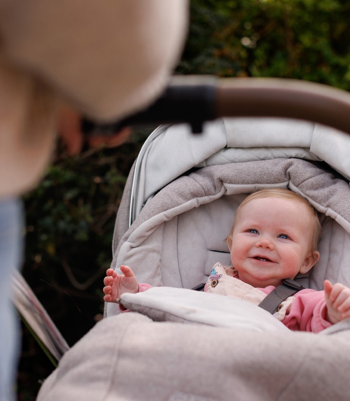 A smiling baby in a stroller looks up at an adult, surrounded by green foliage in the background