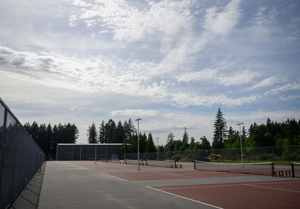 Empty tennis courts under a partly cloudy sky, surrounded by trees and fenced in, with a distant view of the horizon.