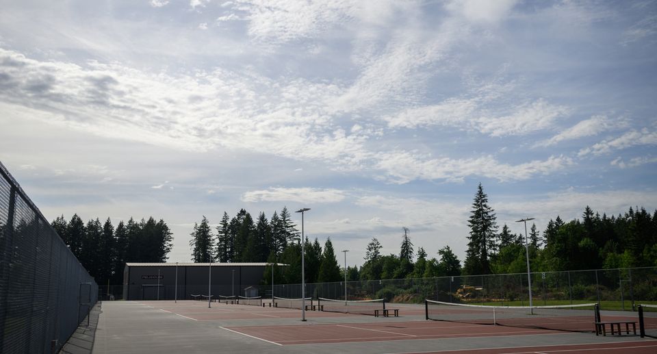 Empty tennis courts under a partly cloudy sky, surrounded by trees and fenced in, with a distant view of the horizon.