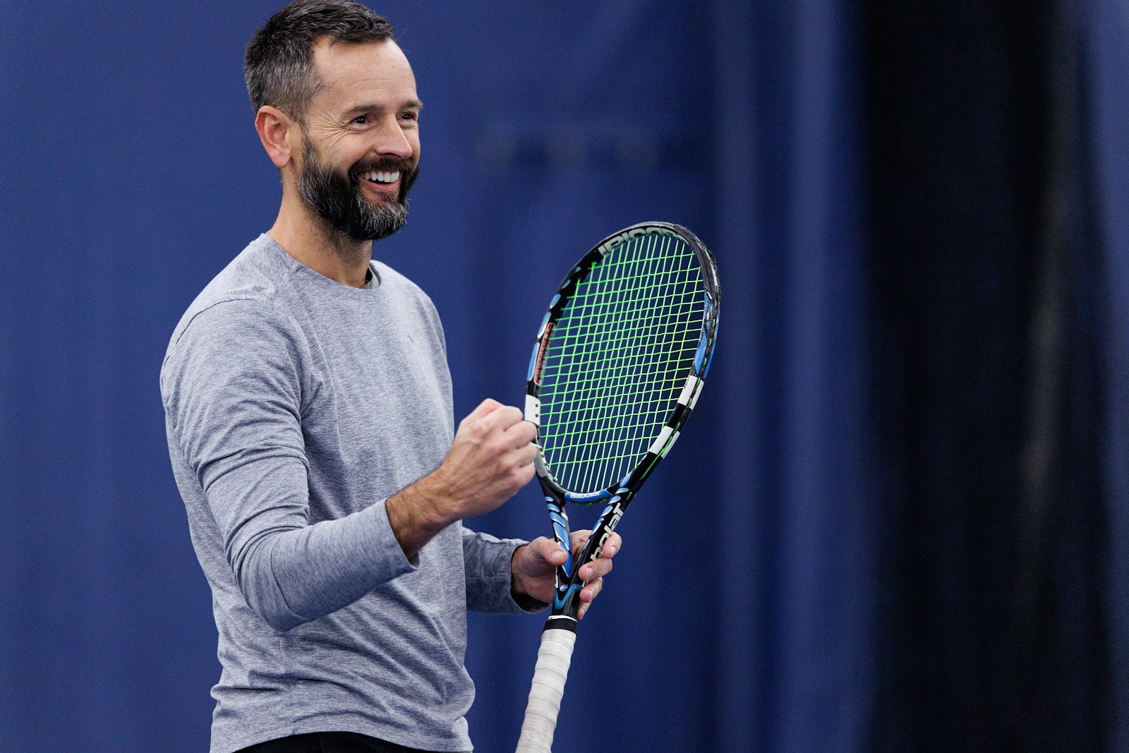 A man in a gray shirt smiles and clenches his fist while holding a tennis racket on an indoor court with a blue background.