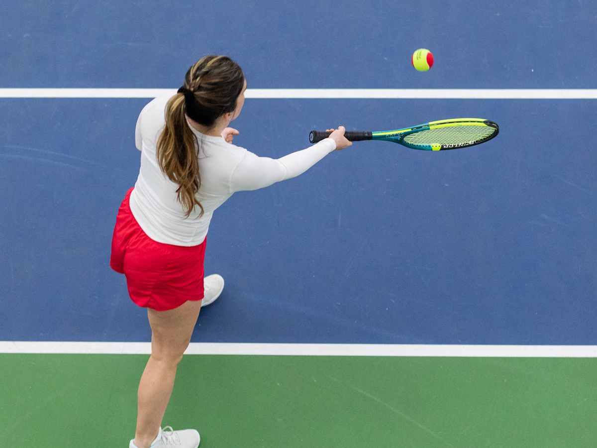 A woman in a white top and red skirt plays tennis on a blue and green court, about to hit a ball with her racket.