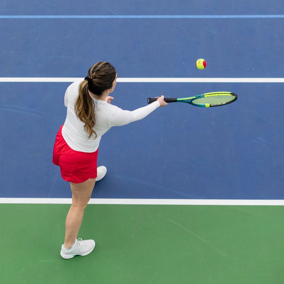 A woman in a white top and red skirt plays tennis on a blue and green court, about to hit a ball with her racket.