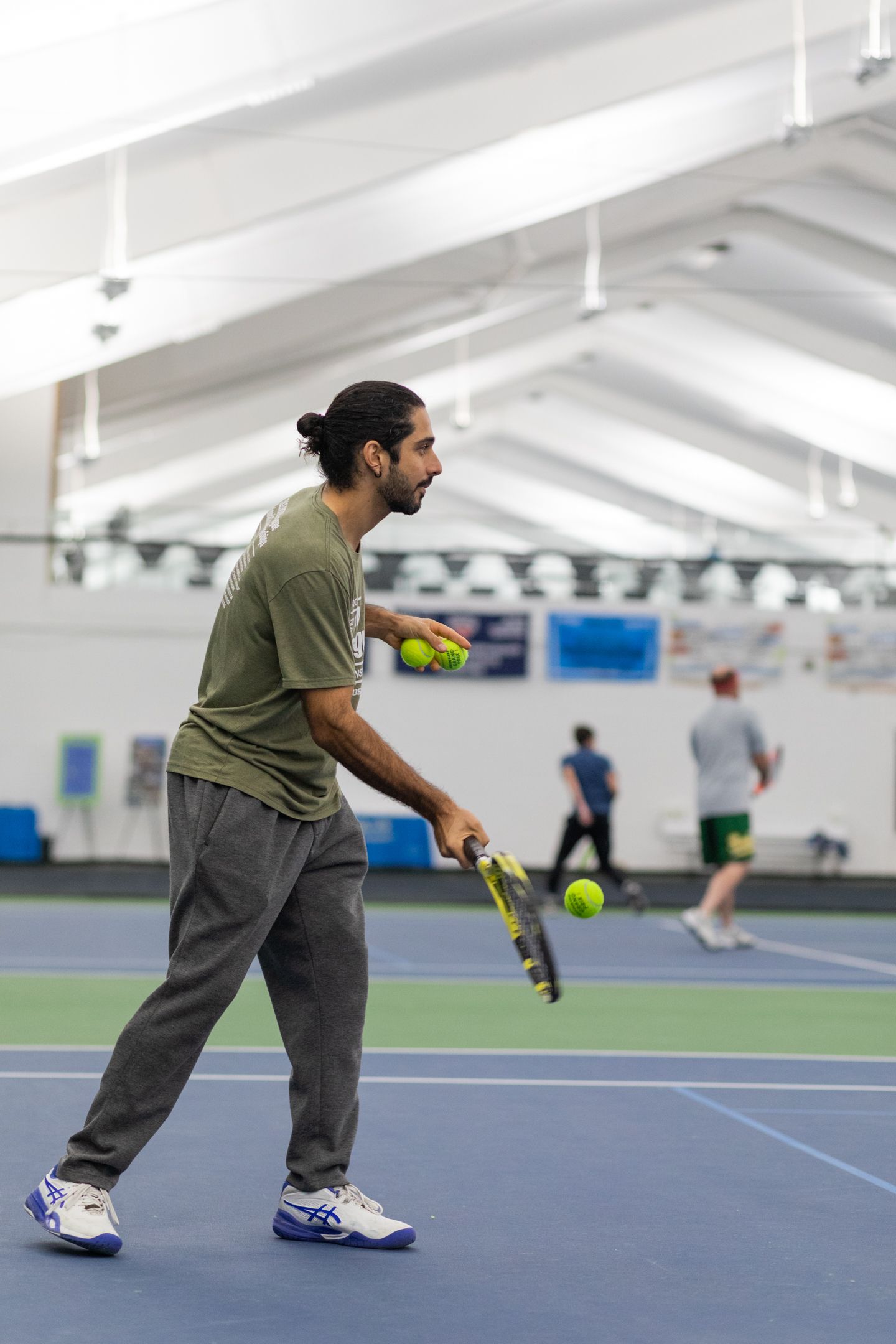 A man in a green shirt and gray pants teaches tennis on an indoor court, holding multiple balls and a racket, with others in the background.