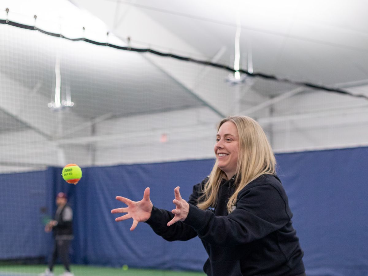 A woman in a black sports attire attempts to catch a tennis ball on an indoor court, smiling with arms outstretched.
