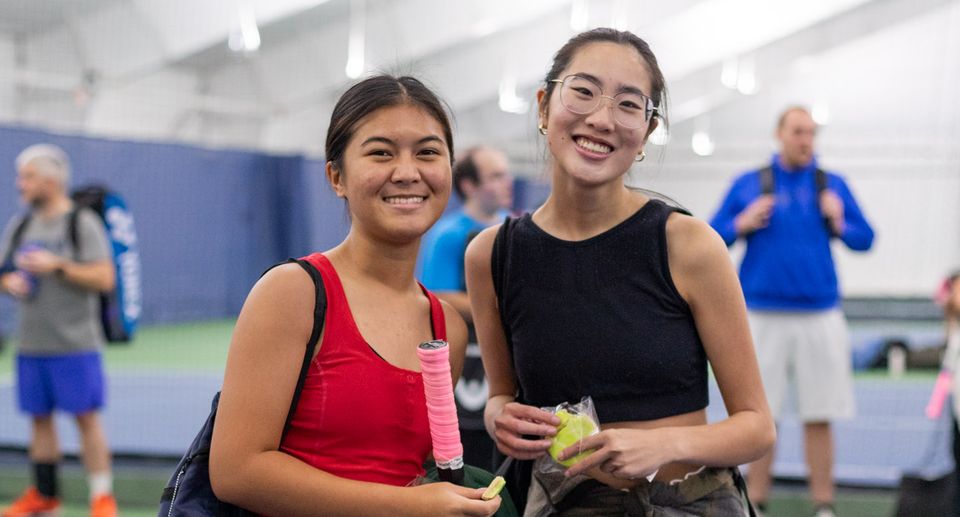 Two women smiling on an indoor tennis court. One holds a racket; both wear athletic attire. Other players are in the background.