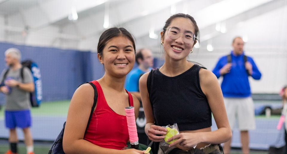 Two women smiling on an indoor tennis court. One holds a racket; both wear athletic attire. Other players are in the background.
