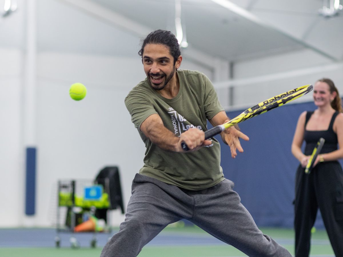 Man playing tennis indoors, focusing on hitting the ball. Another person and tennis equipment are visible in the background.
