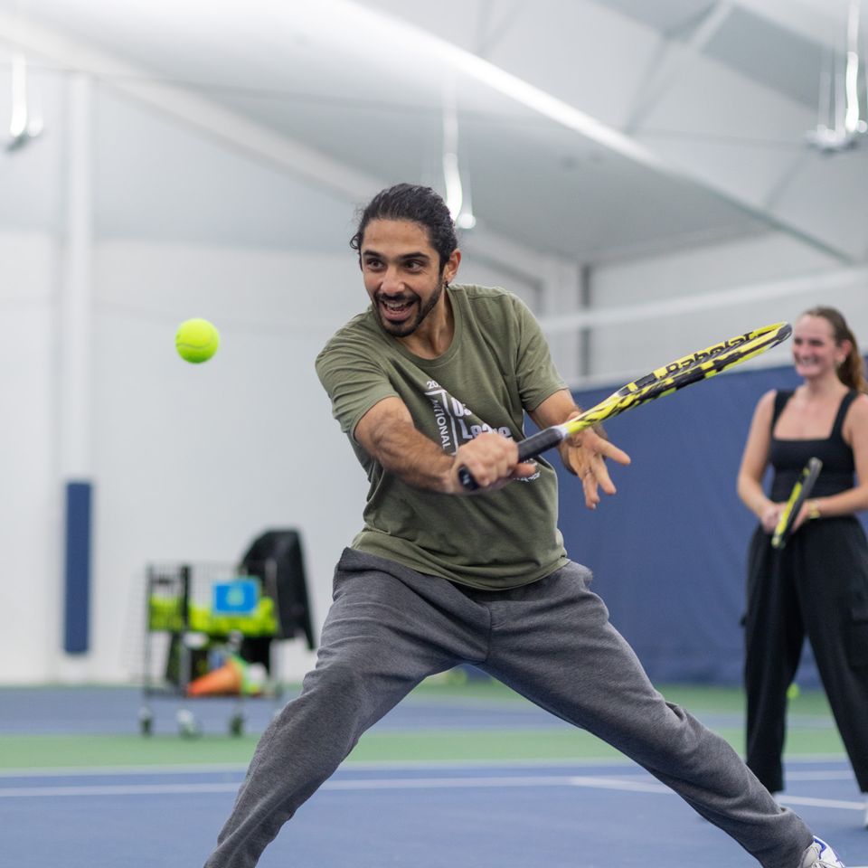 Man playing tennis indoors, focusing on hitting the ball. Another person and tennis equipment are visible in the background.