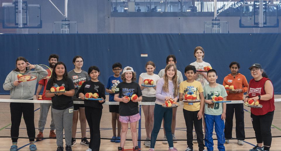 Group of children and two adults holding tennis rackets and tennis balls, standing on an indoor tennis court.