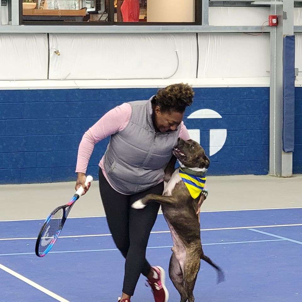 Woman on indoor tennis court hugging a service dog wearing a colorful bandana, holding a tennis racket, both appearing joyful.