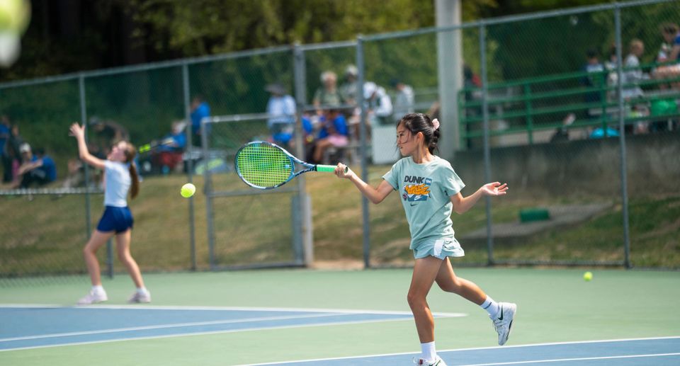 Young girls playing tennis on an outdoor court; one returning a ball with a forehand swing, while another prepares to serve in the background.