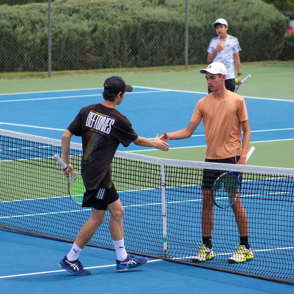 Two tennis players shake hands at the net after a match, with a third player observing in the background on a blue court.
